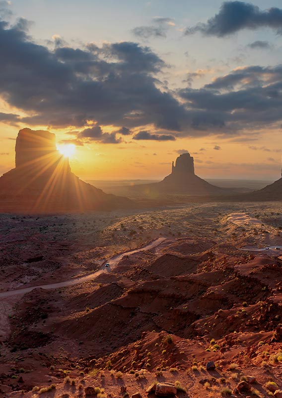 A desert landscape with three tall rock formations rising from the earth, with the sun behind.