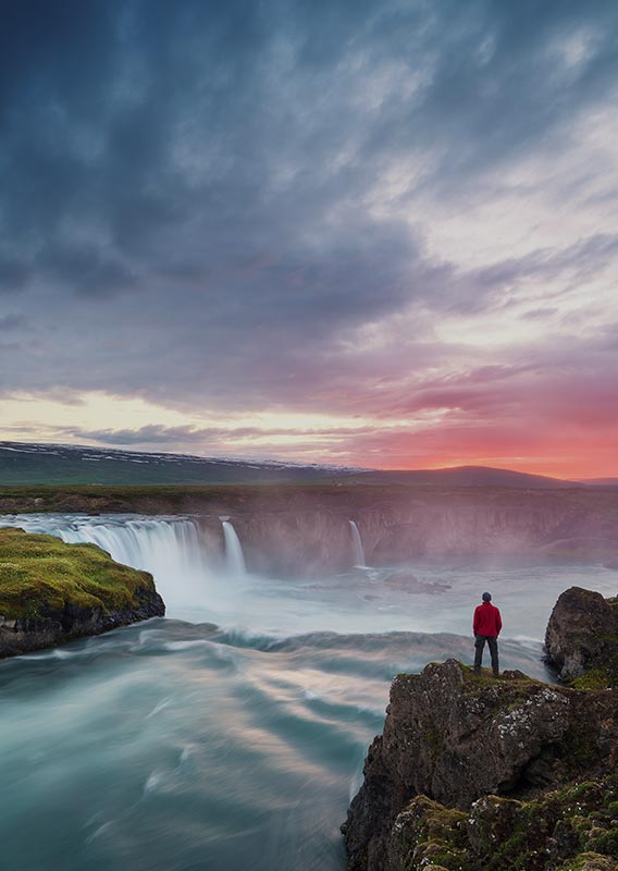 A person stands at a cliff overlooking a river and wide waterfall.